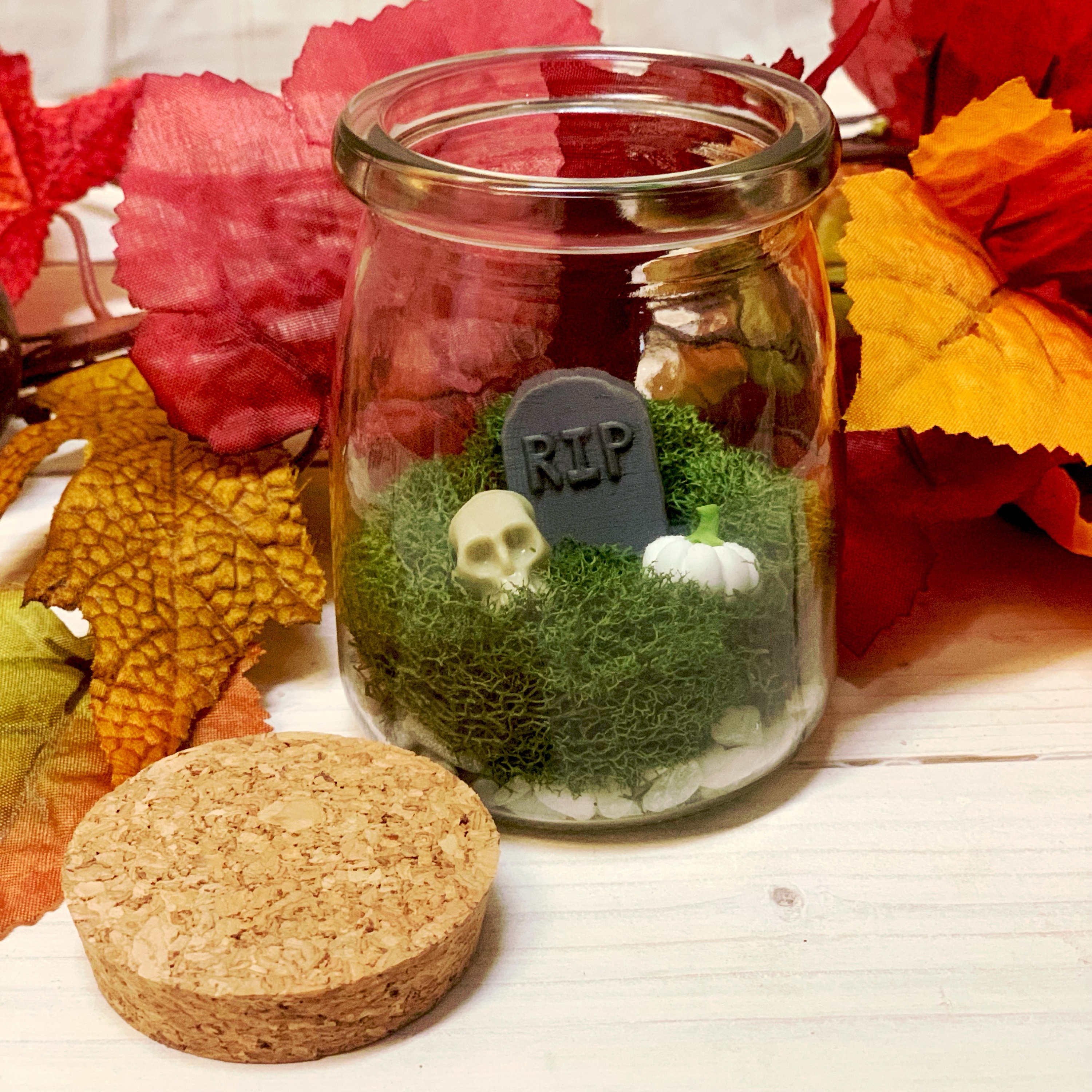 Photo of miniature cemetary terrarium containing headstone, skull, and pumpkin on top of moss.